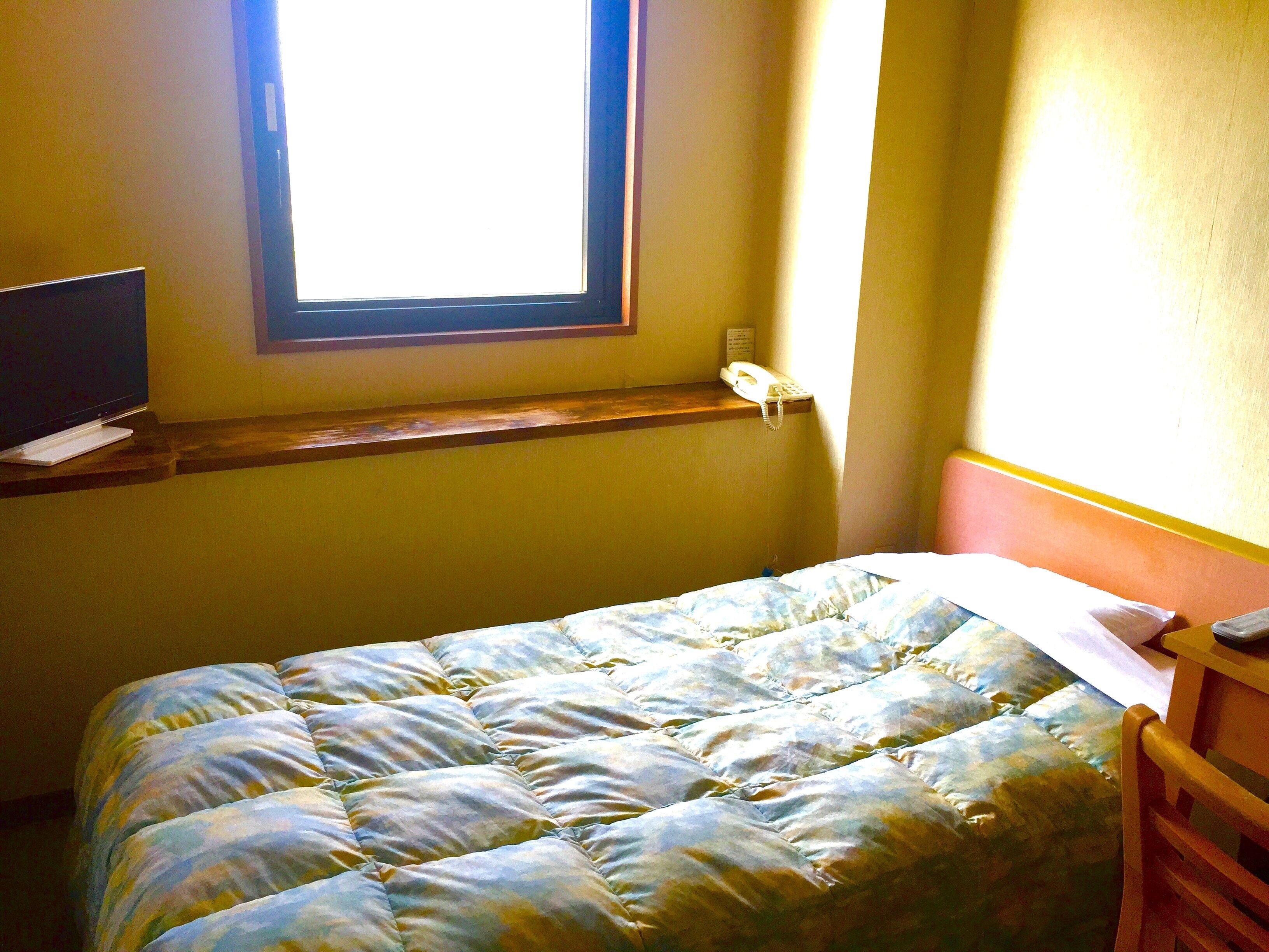 A room with a bed, a television on a stand, a window, and a telephone on a shelf.