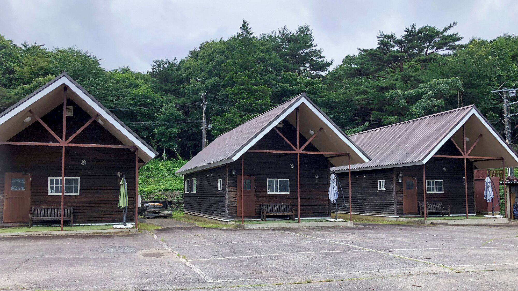 Drei Holzhütten mit Satteldächern, ein gepflasterter Parkplatz und ein Hintergrund aus dichtem Wald.