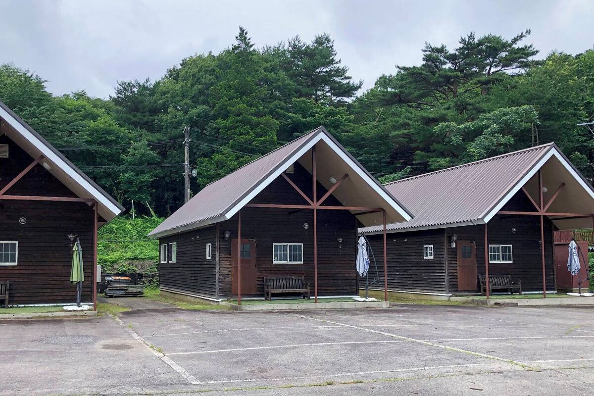 Three wooden cabins with pitched roofs, a paved parking area, and a backdrop of dense trees.
