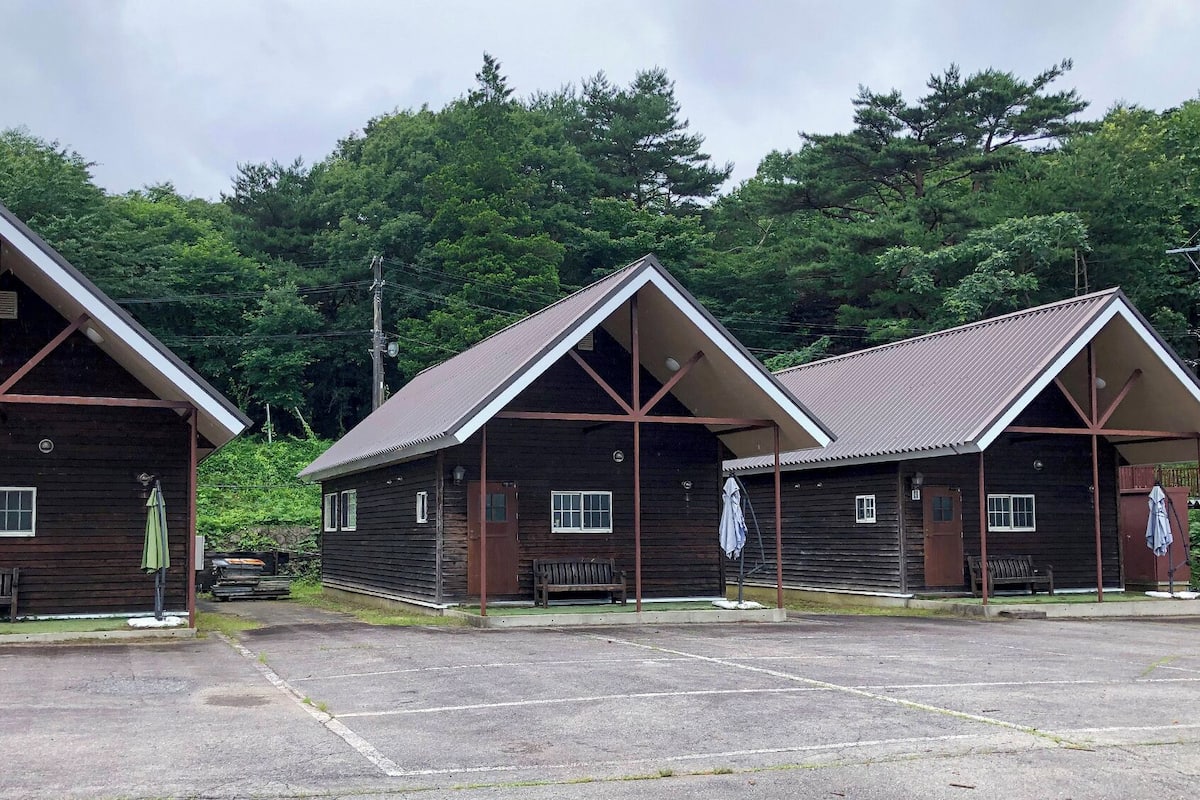 Three wooden cabins with pitched roofs, a paved parking area, and a backdrop of dense trees.
