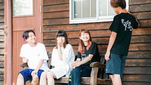 Four people sitting on a wooden bench outside a cabin with a window above them.