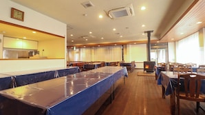 A large dining hall with long tables, a kitchen area, and a fireplace.