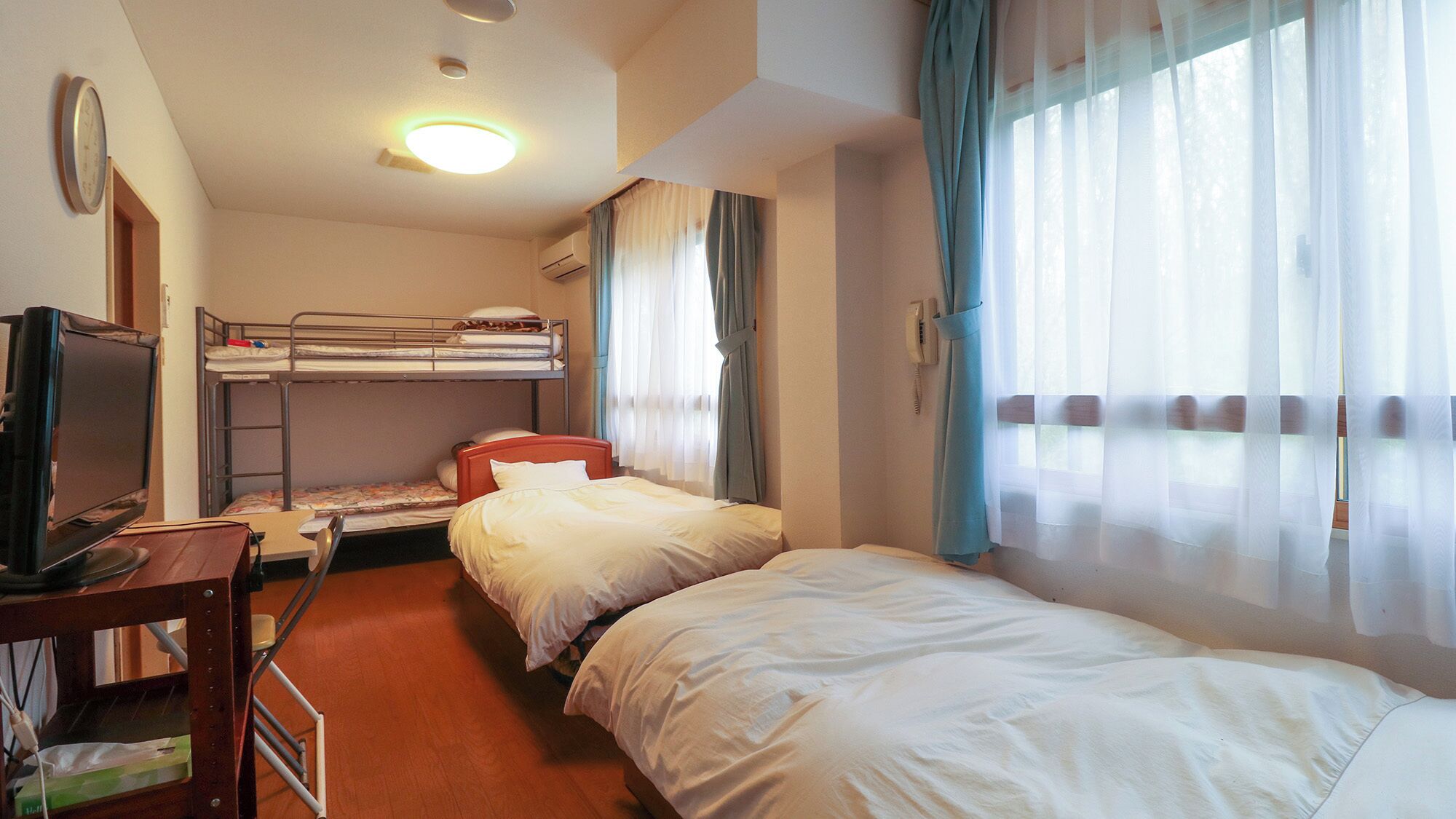 Ein Hotelzimmer mit Etagenbetten, einem Schreibtisch mit Computer, einem Telefon und einem Fenster mit Vorhängen.