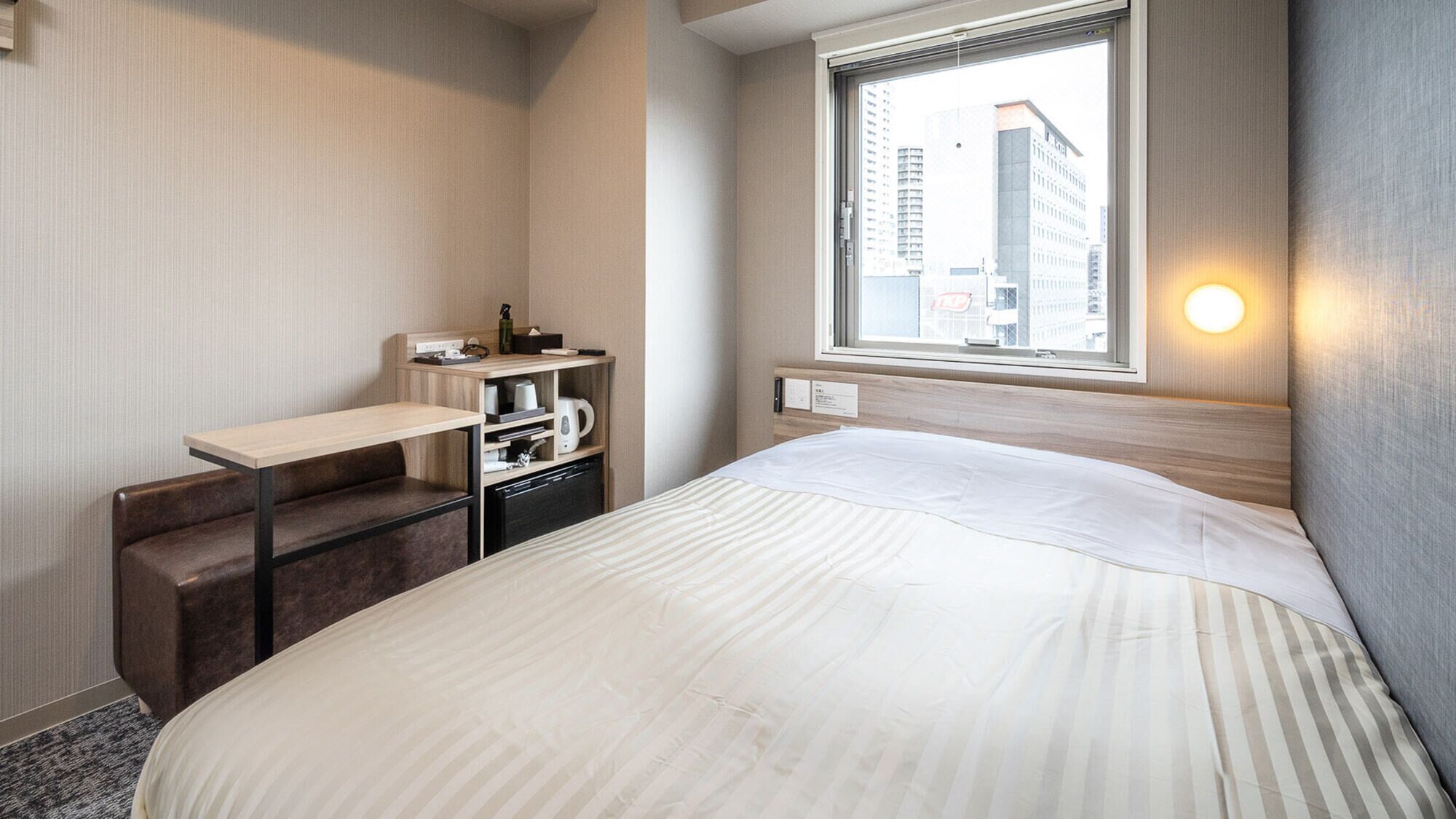 Quarto de hotel com cama, escrivaninha, cadeira, pequena cozinha e janela com vista para a cidade.