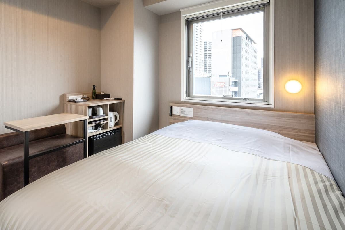 A hotel room with a bed, a desk, a chair, a small kitchenette, and a window with a city view.