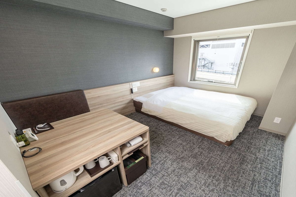A compact hotel room with a bed, a small desk, a chair, a window, and a television.