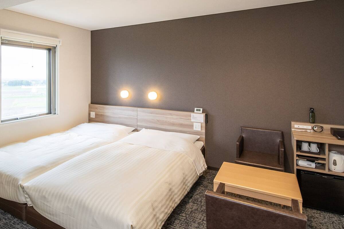 A hotel room with a bed, a chair, a small table, a shelf with a kettle, and a window.