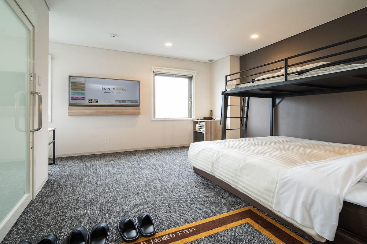 A hotel room with a bunk bed, a flat-screen TV, a window, and a shoe rack.