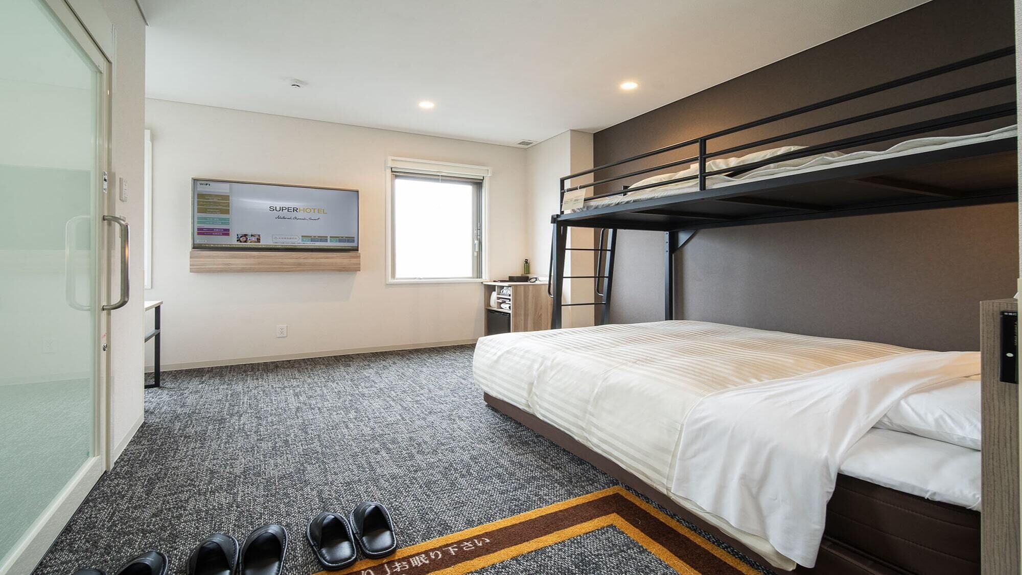 Quarto de hotel com uma cama beliche, televisão de tela plana, janela e sapateira com sapatos.