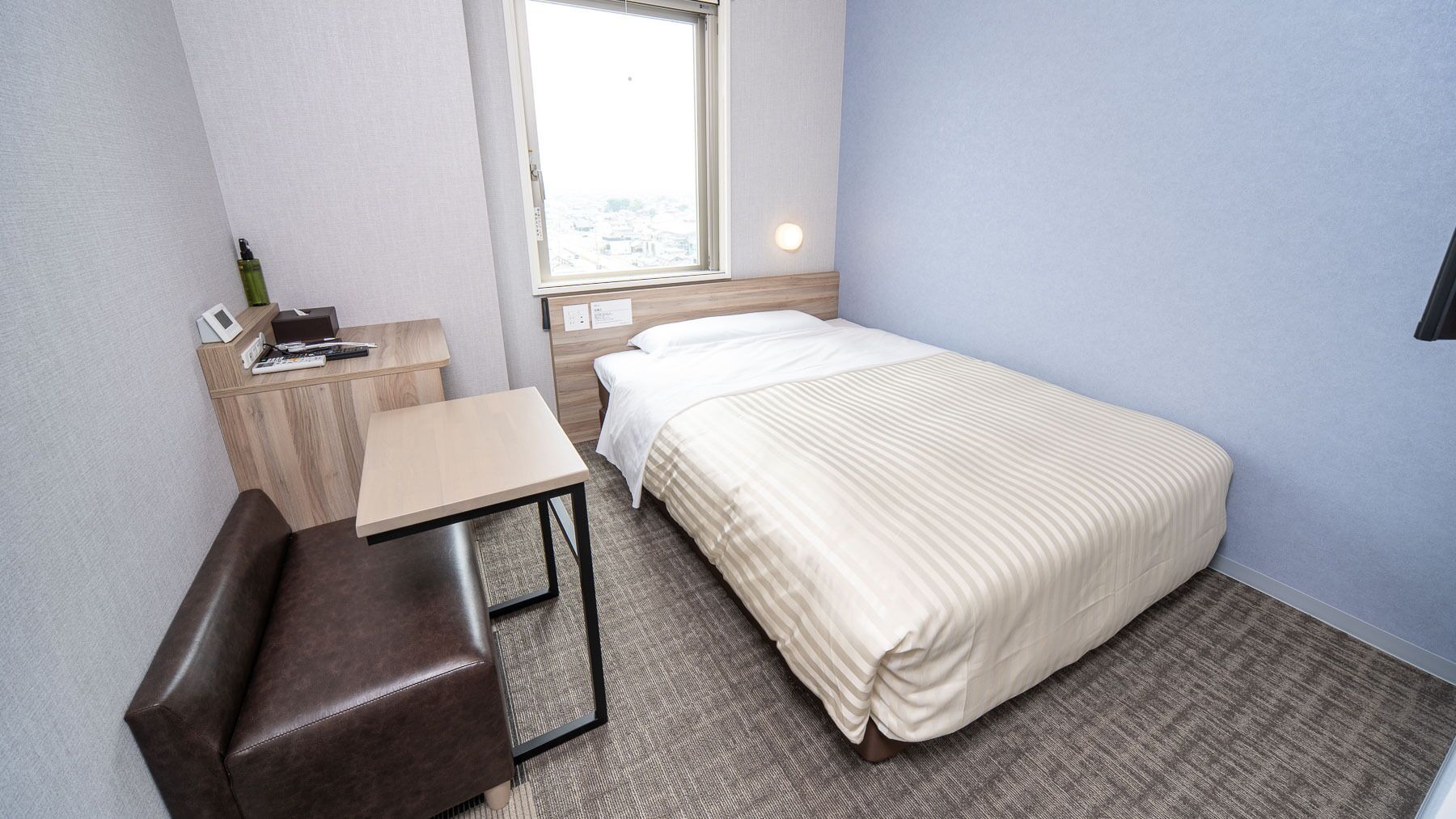 Quarto de hotel pequeno e bem organizado, com uma cama, uma escrivaninha, uma cadeira e uma janela.