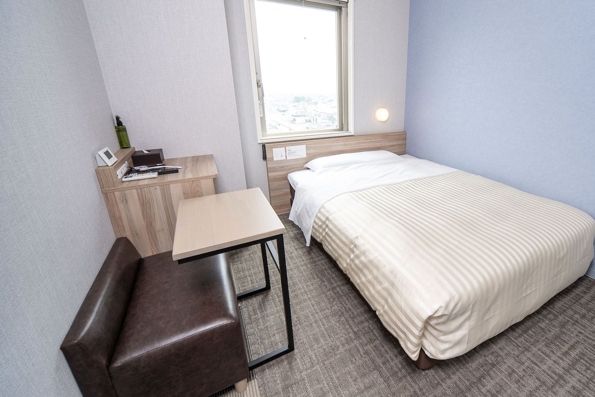 A small, neatly arranged hotel room with a bed, a desk, a chair, and a window.