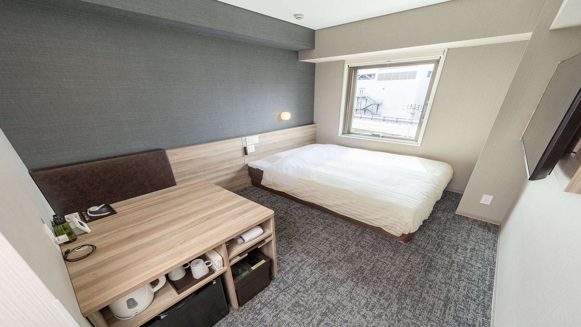 Quarto de hotel pequeno com cama, escrivaninha, cadeira, janela e televisão.