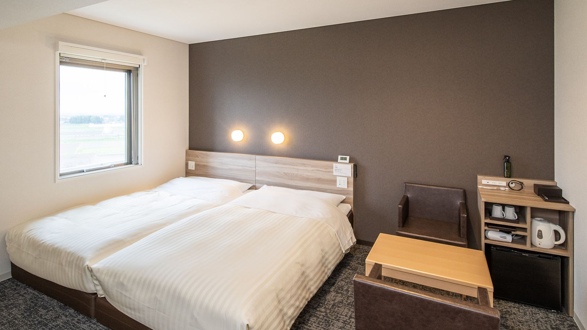 Quarto de hotel com cama, uma cadeira, uma mesinha, uma prateleira com chaleira e uma janela.