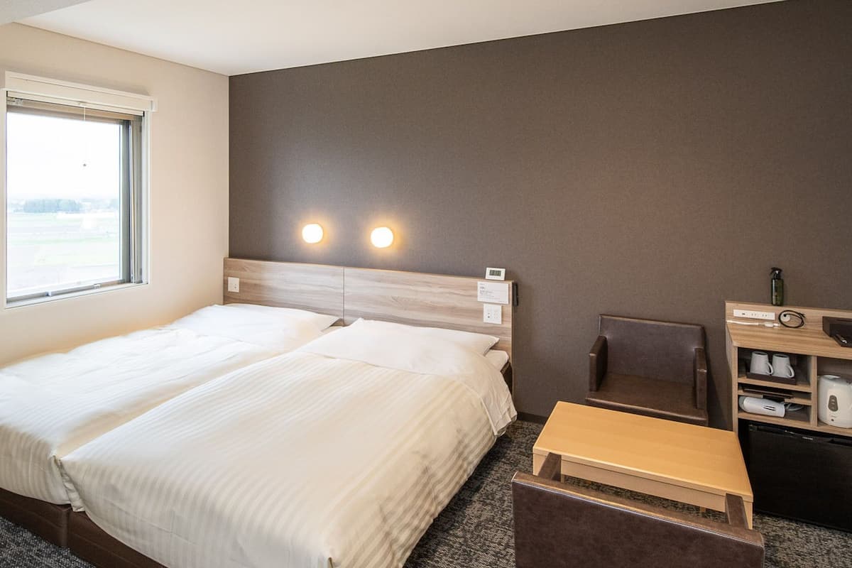 A hotel room with a bed, a chair, a small table, a shelf with a kettle, and a window.