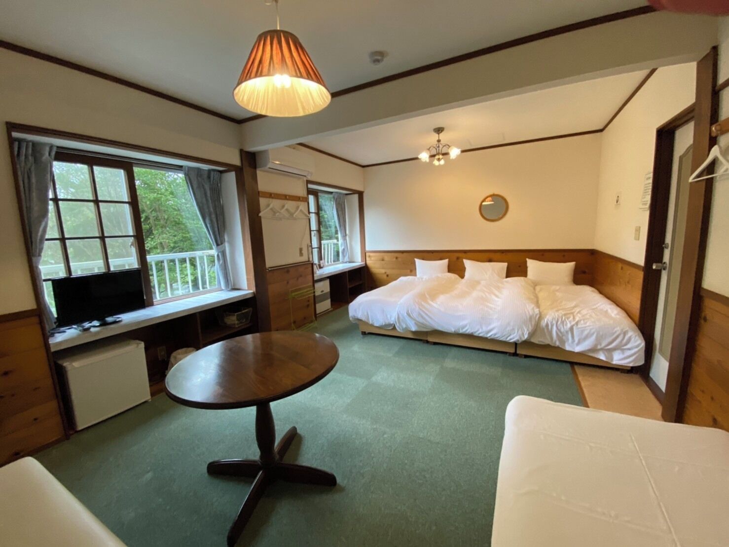 A bedroom with a bed, a round table, a TV, and a window with a view of greenery.