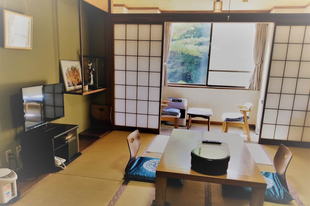 Een traditionele Japanse kamer met tatami vloerbedekking, een lage tafel met een zwarte dienblad, een televisie en een raam met uitzicht op groen.