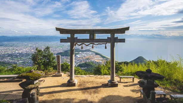 Een traditionele Japanse torii poort met zicht op een kuststad en bergen op de achtergrond.