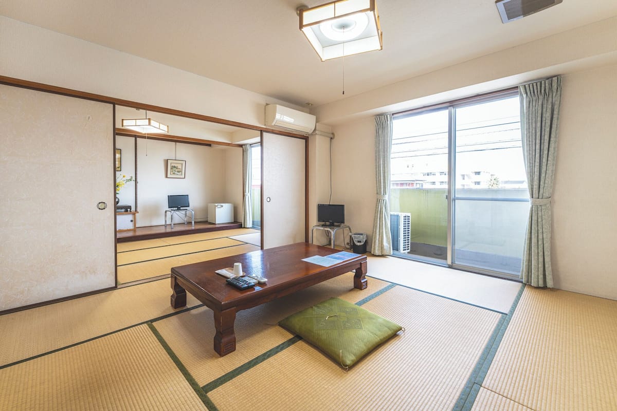 A traditional Japanese room with tatami flooring, a wooden table, a green cushion, a television, and a sliding glass door to the outside.