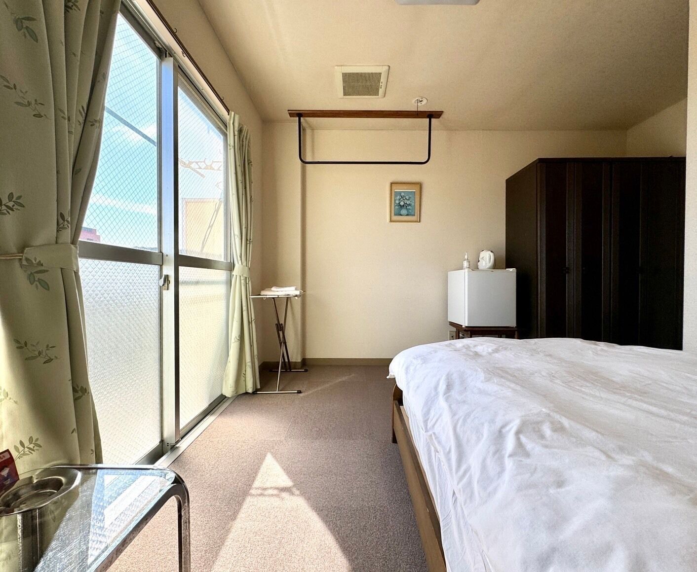 A hotel room with a bed, a small table, a window with curtains, a wall-mounted radiator, and a wardrobe.