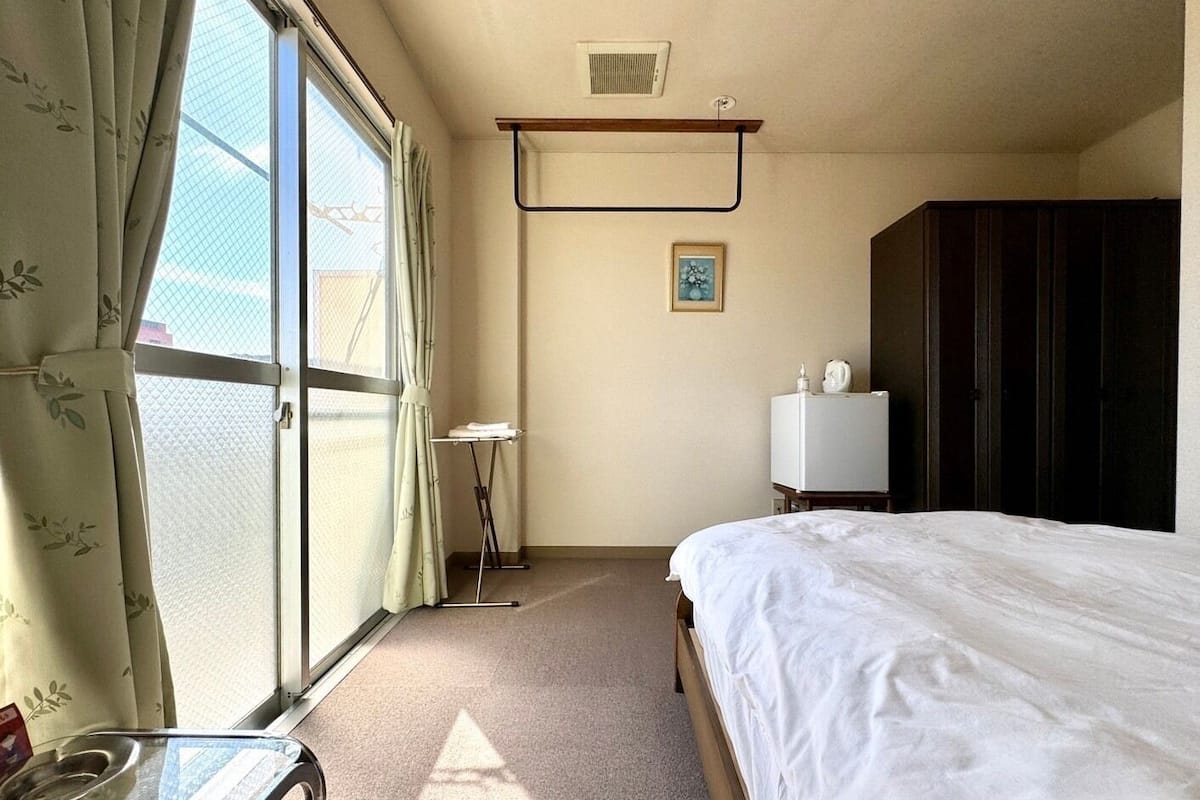A hotel room with a bed, a small table, a window with curtains, a wall-mounted radiator, and a wardrobe.