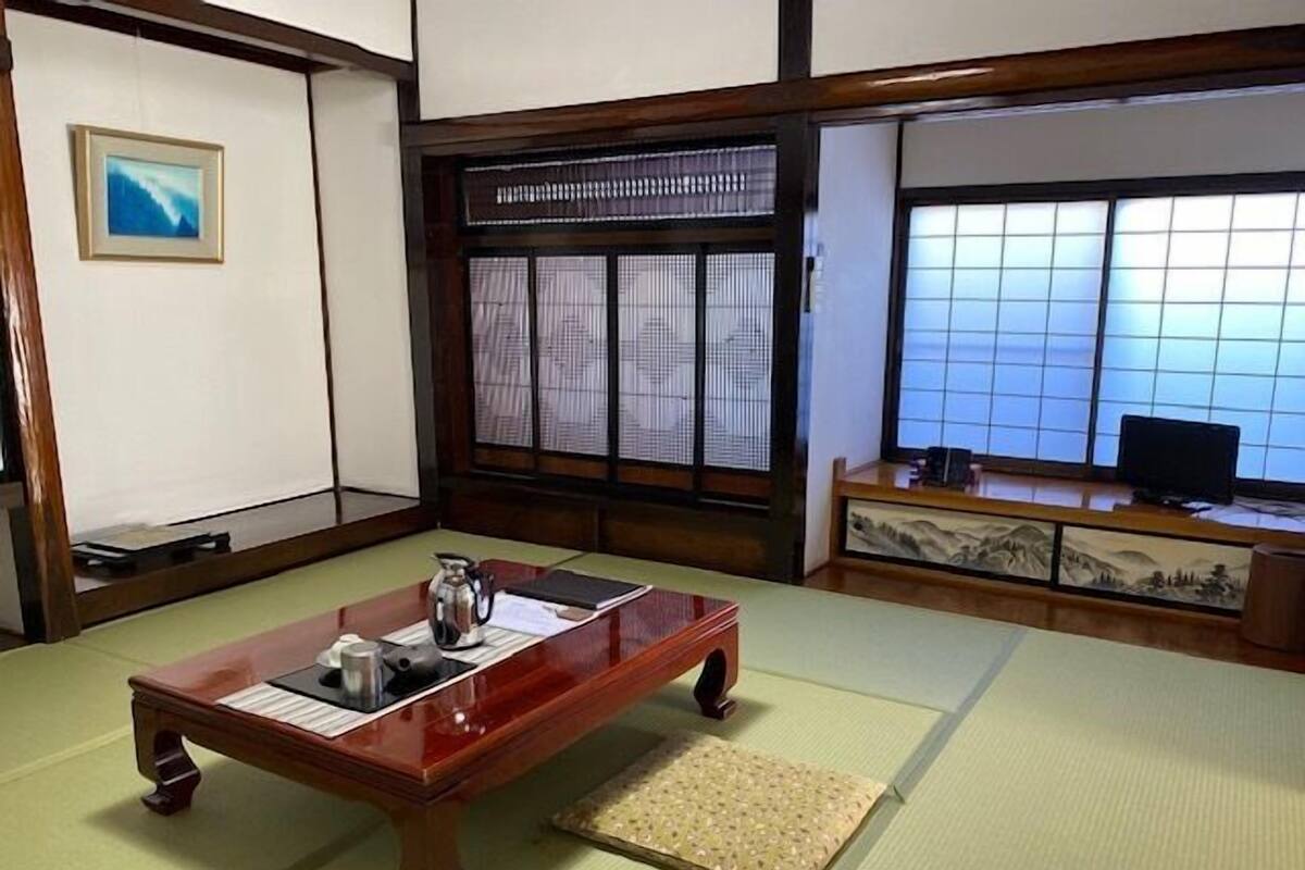 Une pièce de style japonais traditionnel, avec un sol recouvert de tatamis, une petite table en bois sur laquelle se trouve une bouilloire, un téléviseur posé sur un meuble en bois et un paravent shoji.
