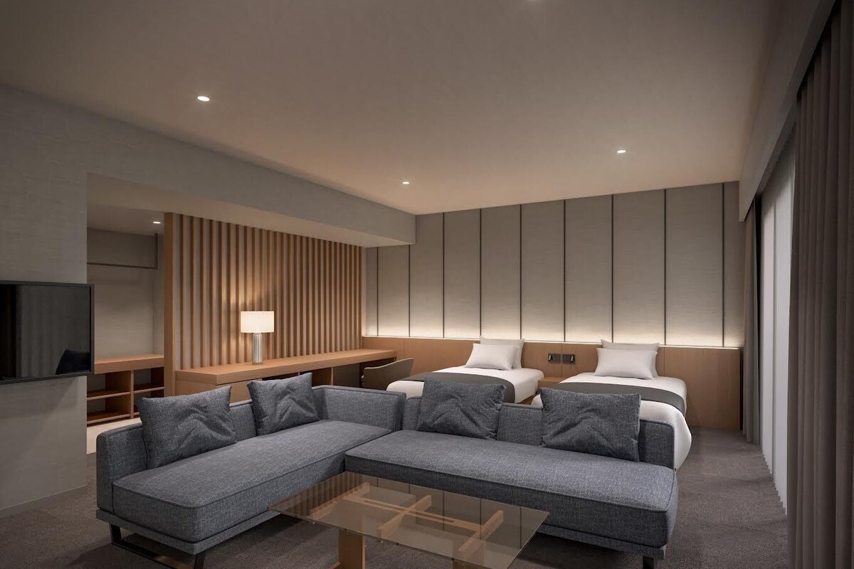 A modern hotel room with a sectional sofa, a glass coffee table, a TV, and a bed with headboards.