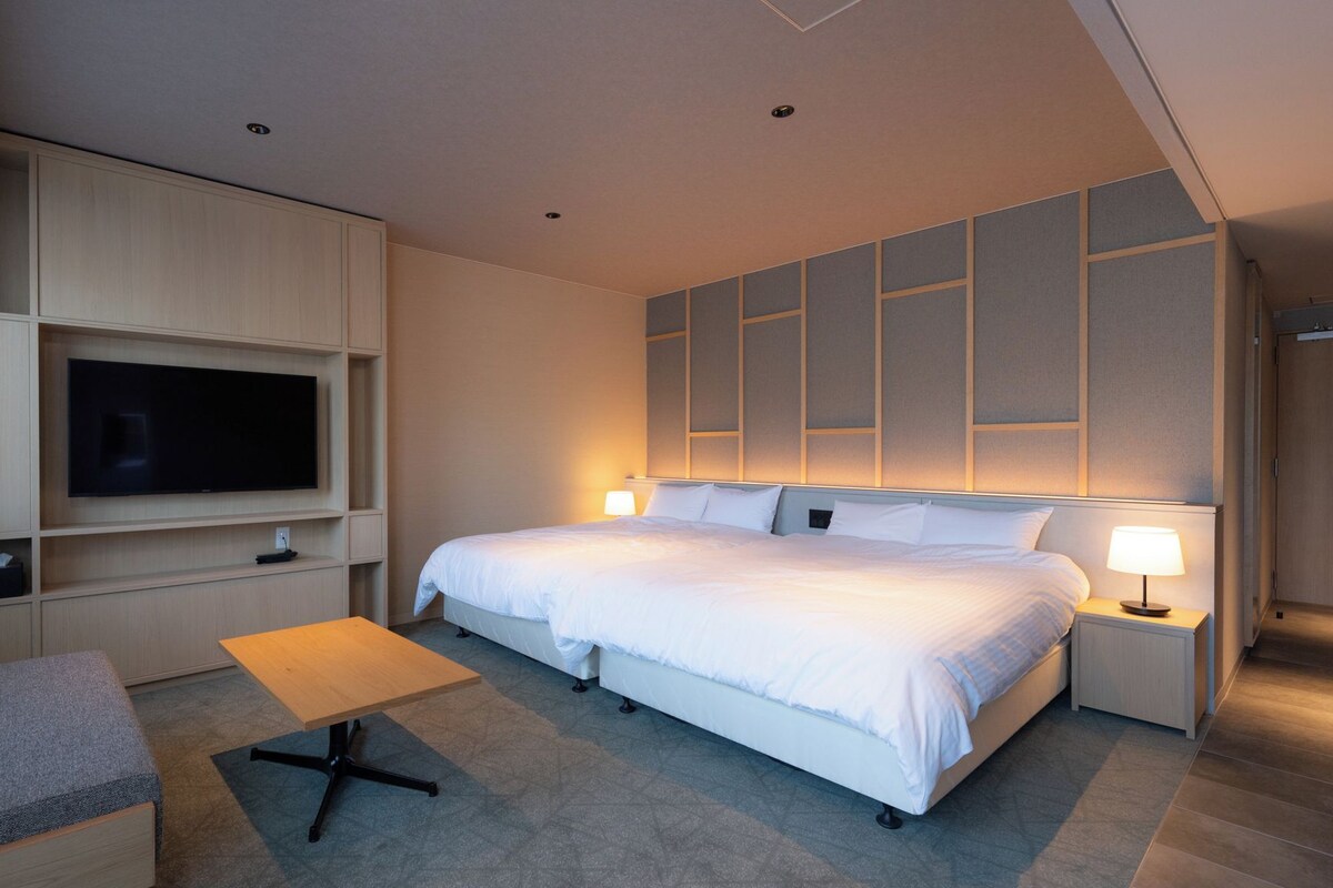 A modern hotel room with a large bed, a TV mounted on the wall, a small table, and a sofa.