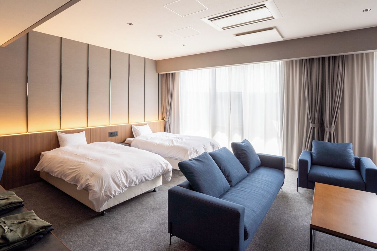 A hotel room with two beds, a sofa, a chair, and a table. There is a window with curtains and a view of the outside.