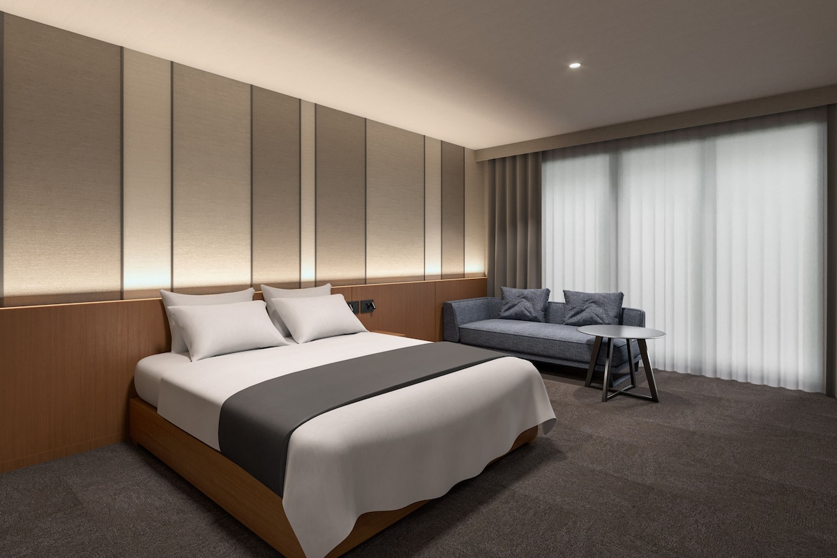 A modern hotel room with a large bed, a sofa, a small table, and a large window with curtains.