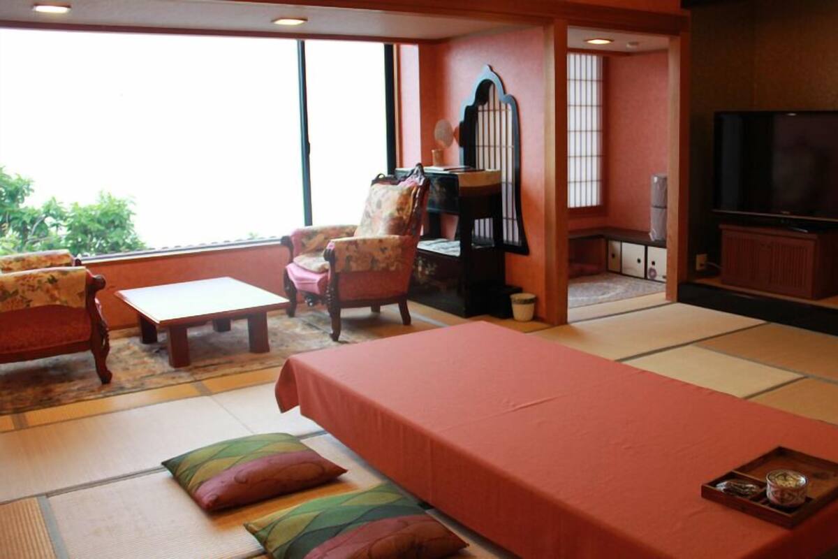 A traditional Japanese-style room with tatami flooring, a low table, a sofa, a chair, a television, and a sliding door leading to another room.