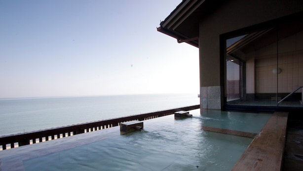 A spa pool with a view of the sea, a wooden deck, and a building with large windows. - Tara Takezaki Onsen Kaijyoukan (Tara)