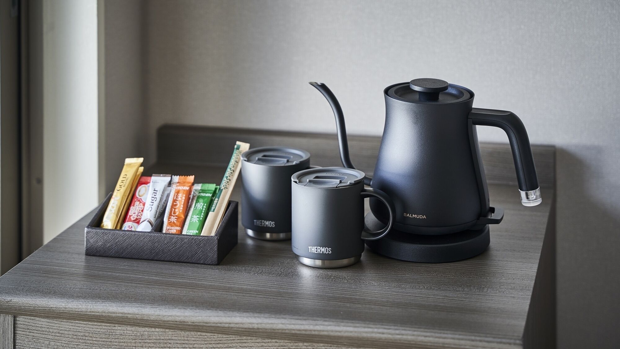 A black electric kettle, two black insulated mugs, and a box with tea bags on a wooden surface.