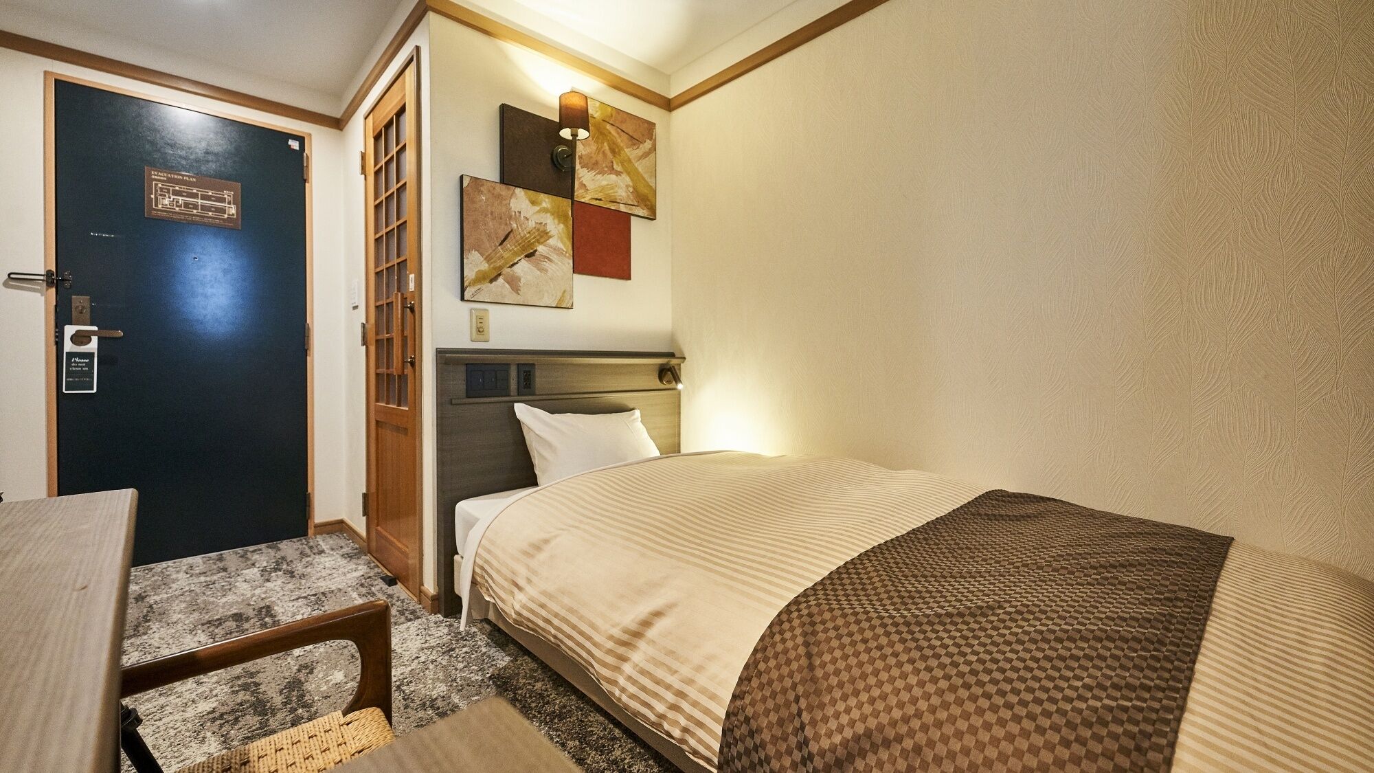 A hotel room with a bed, a desk, a chair, a door, and a wall-mounted artwork.