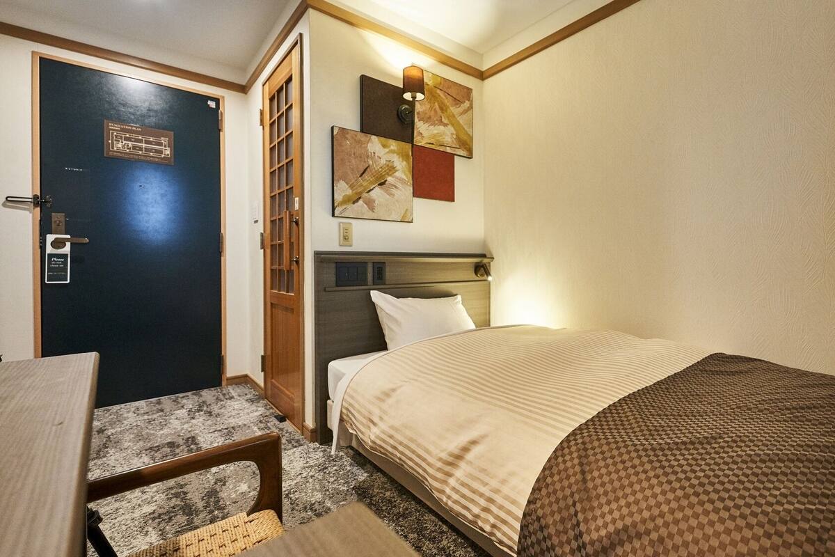 A hotel room with a bed, a desk, a chair, a door, and a wall-mounted artwork.