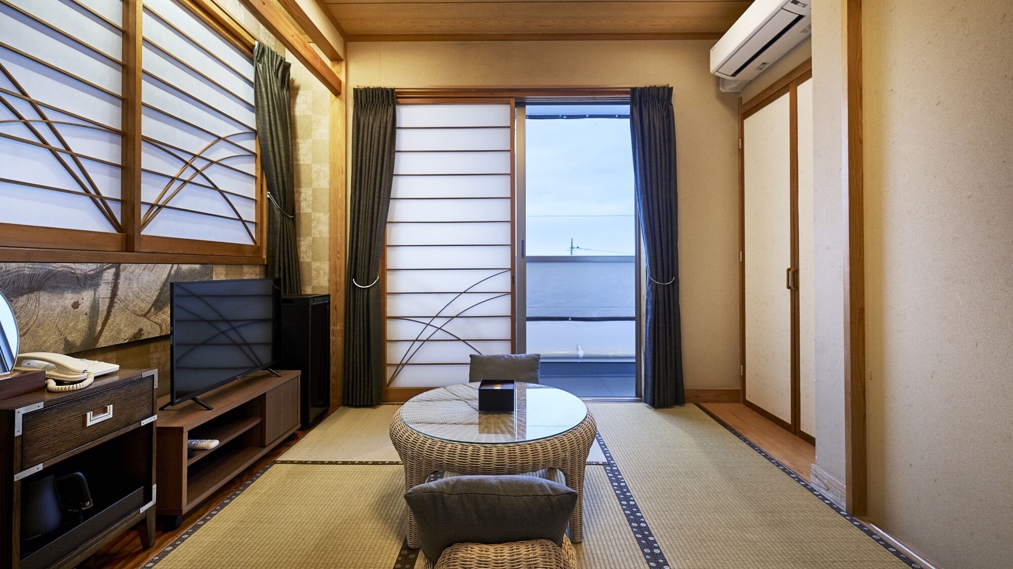 A traditional Japanese room with tatami flooring, a low table, a chair, a television, and a sliding door leading to a balcony.
