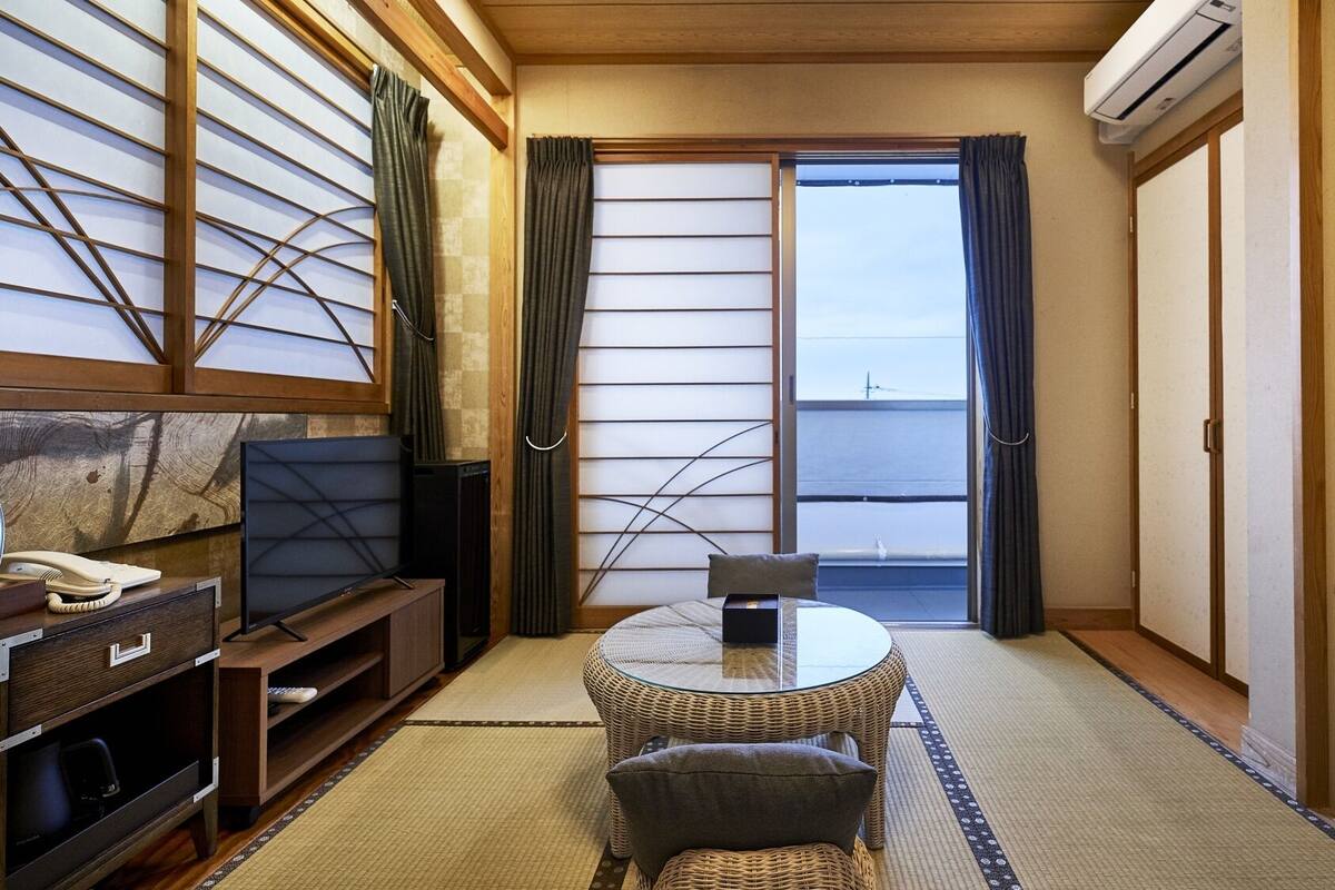 A traditional Japanese room with tatami flooring, a low table, a chair, a television, and a sliding door leading to a balcony.