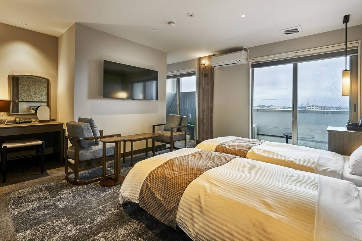 A hotel room with two beds, a desk, a chair, a TV, and a large window with a view.