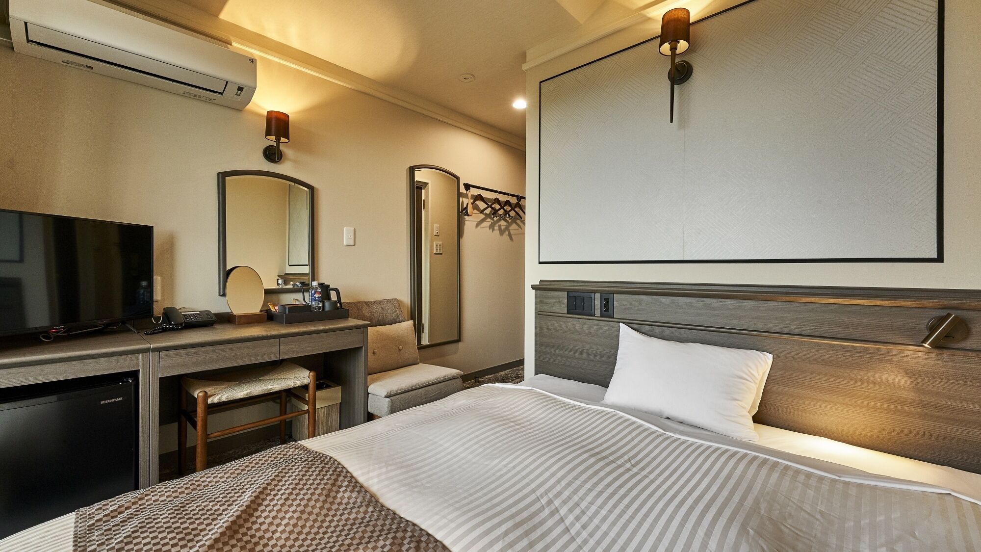 A hotel room with a bed, a desk with a television, a mirror, and a wall-mounted light fixture.