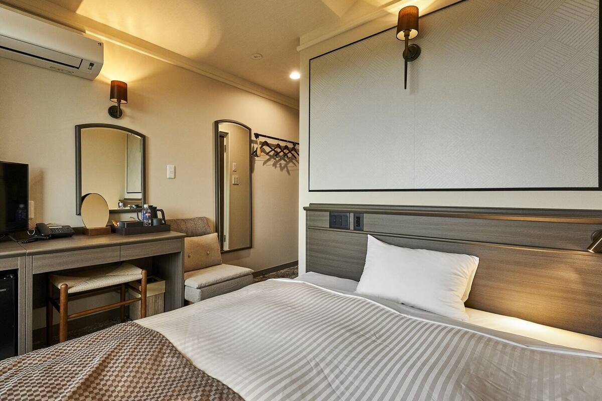 A hotel room with a bed, a desk with a television, a mirror, and a wall-mounted light fixture.