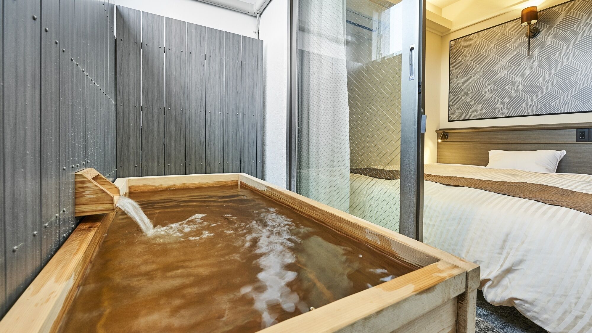 A traditional Japanese onsen with a wooden tub, a bed with a patterned headboard, and a glass partition.