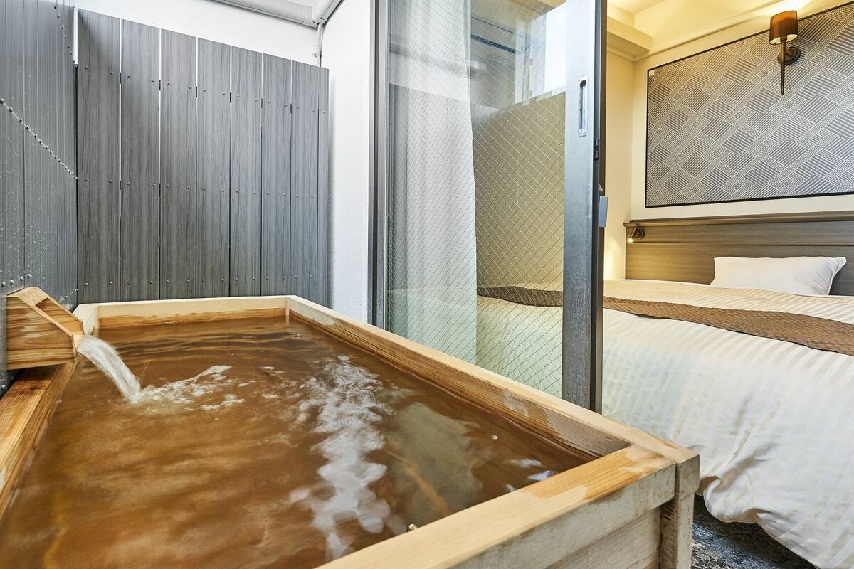 A traditional Japanese onsen with a wooden tub, a bed with a patterned headboard, and a glass partition.