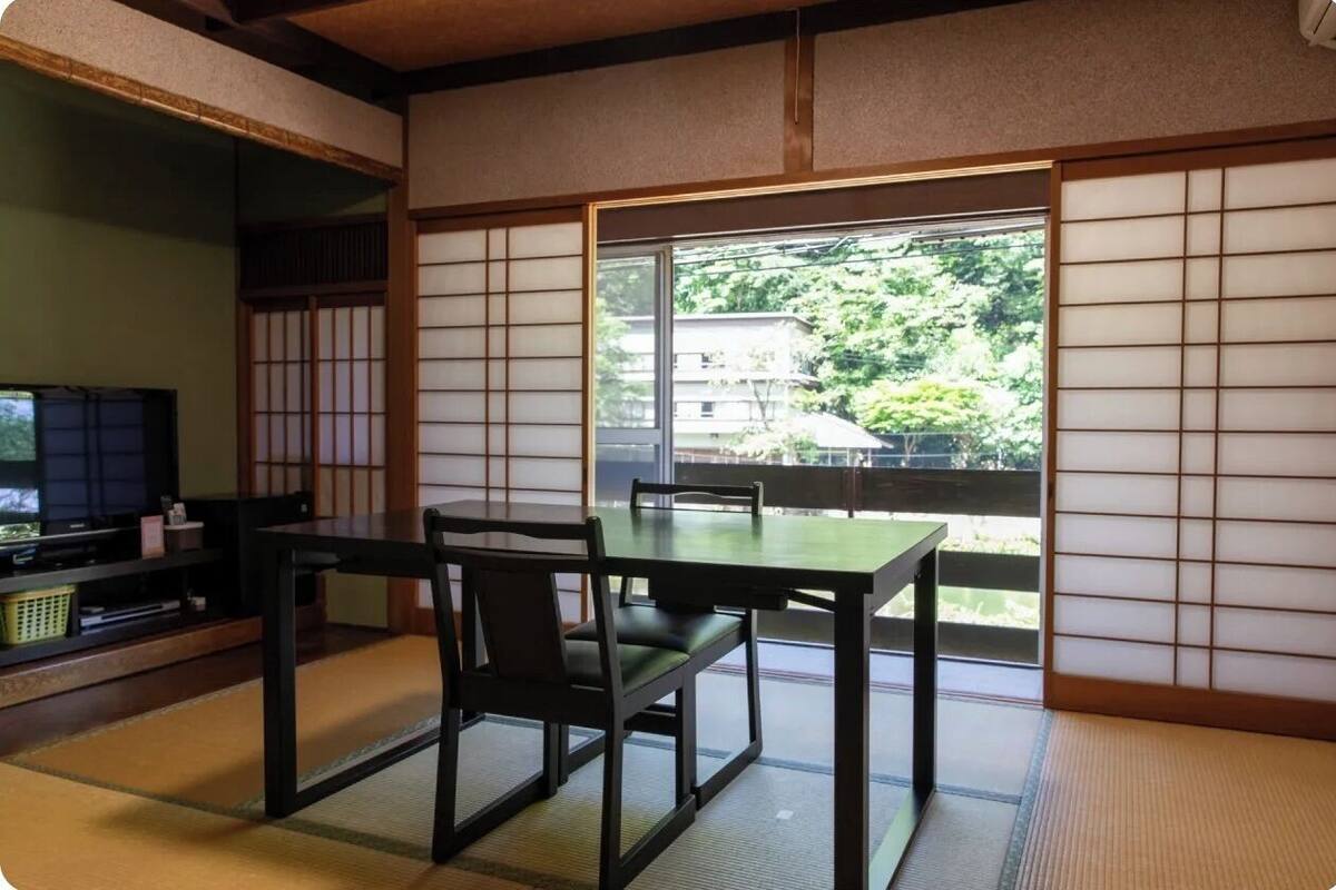 A traditional Japanese room with tatami flooring, a low table with chairs, a television, and a sliding door with a view of greenery.
