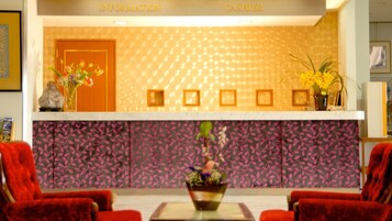 A hotel reception area with a reception desk, a flower vase, and two red chairs.