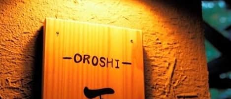 Un letrero de madera con caracteres japoneses y el nombre 'OROSHI' sujeto a una pared texturizada.