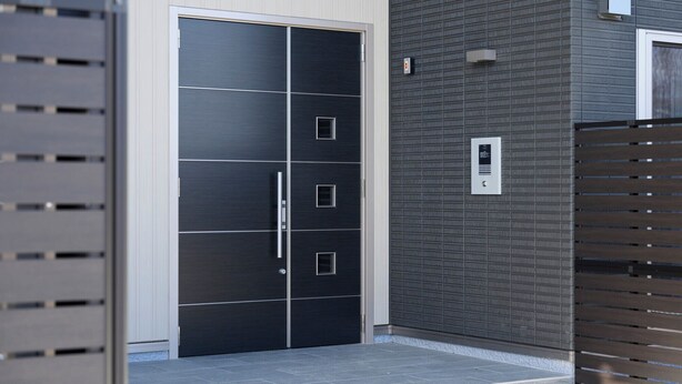 A modern building entrance with a large, dark-colored sliding door, a metal security panel, and a textured wall. - Ties Verde Hotel (Okuma)