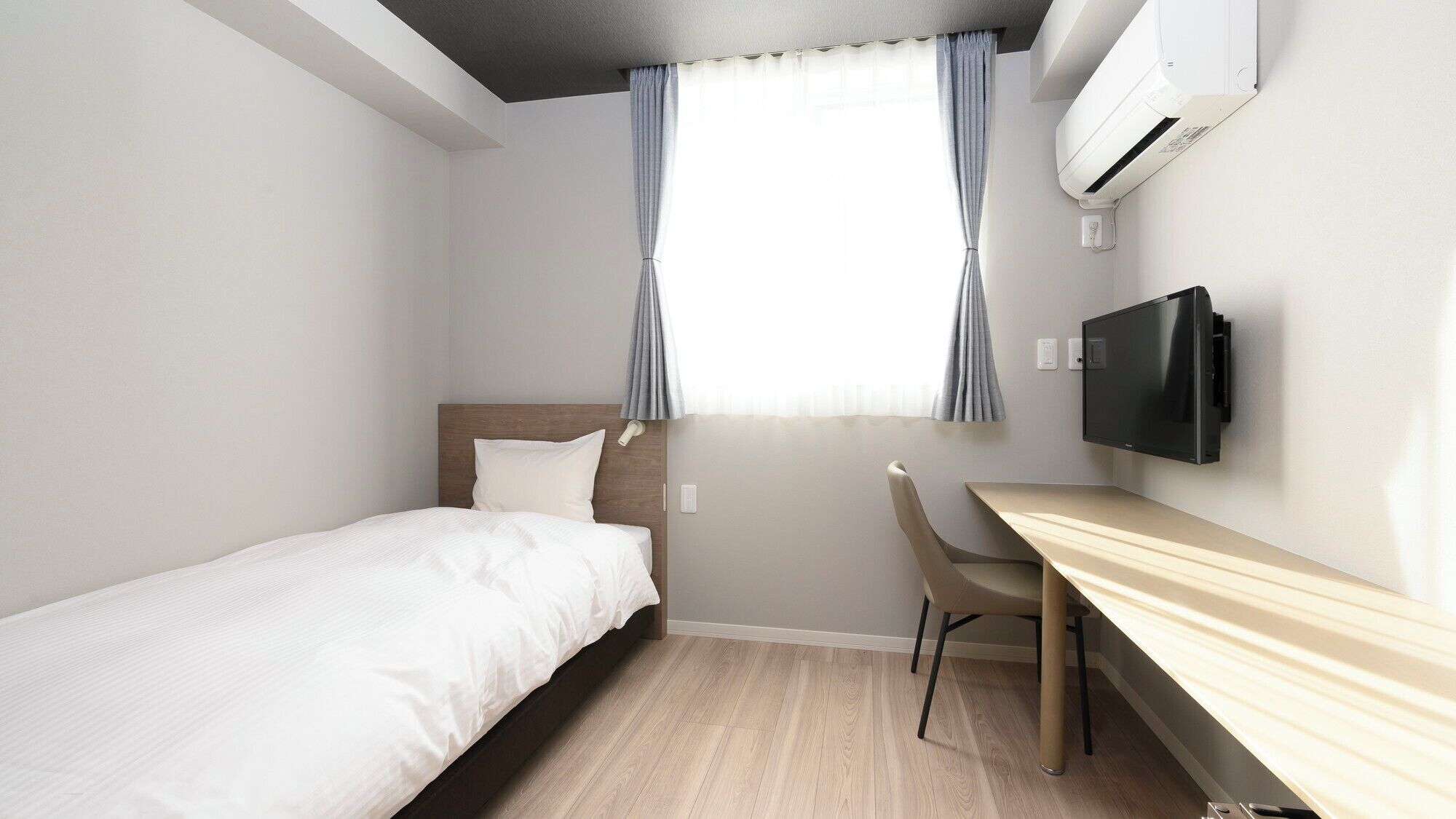 A hotel room with a bed, a desk, a chair, a TV, and an air conditioning unit.