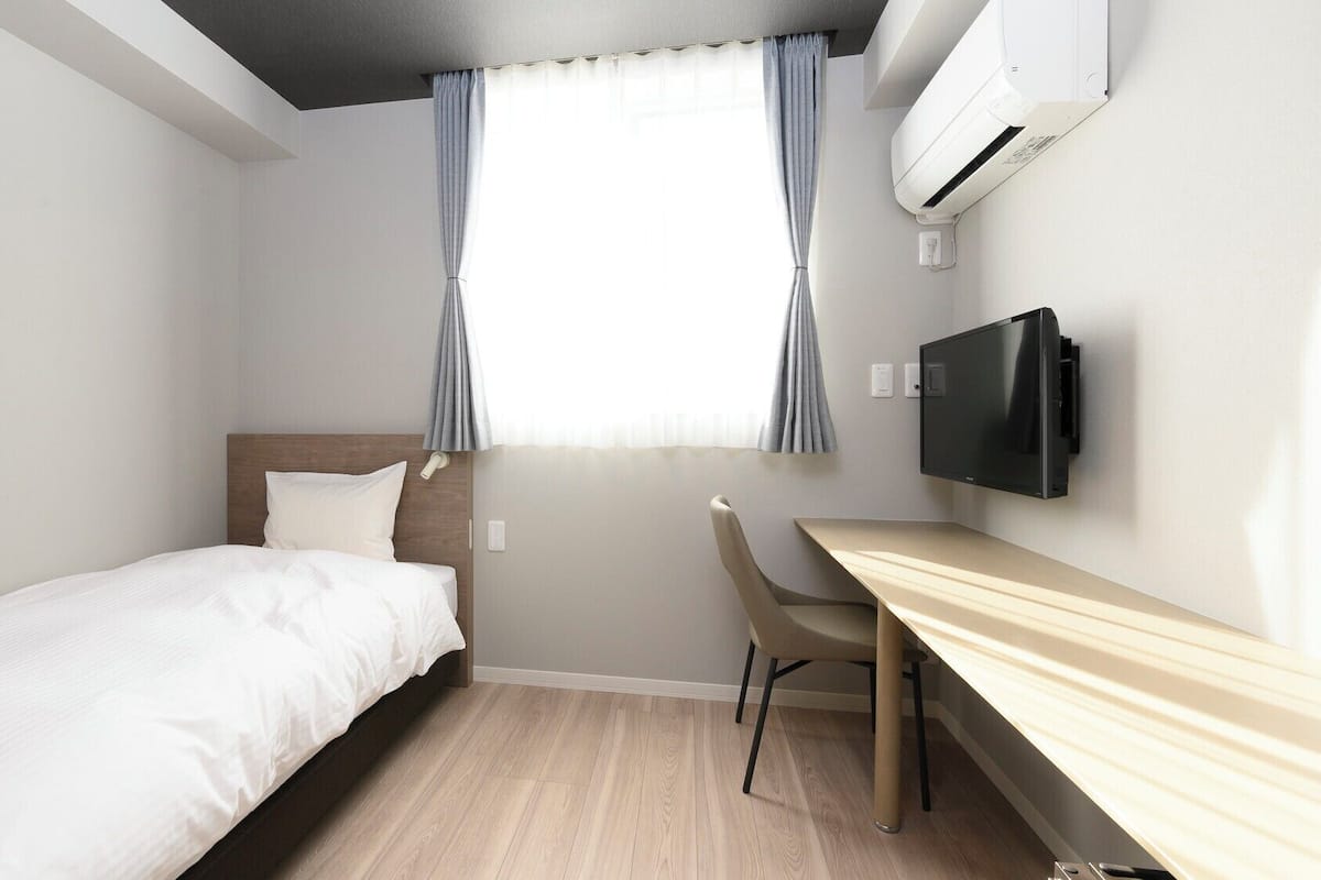 A hotel room with a bed, a desk, a chair, a TV, and an air conditioning unit.