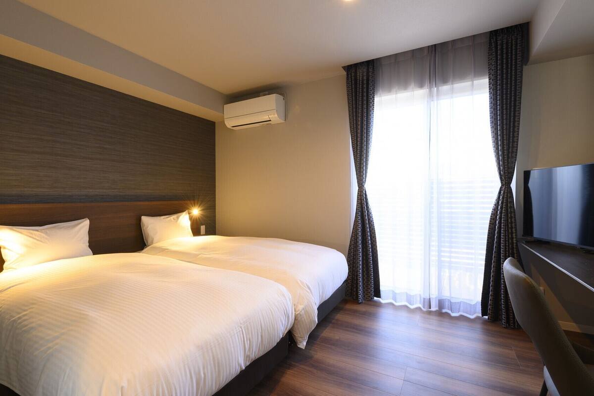 Habitación de hotel con dos camas, un televisor de pantalla plana, un equipo de aire acondicionado y un ventanal con cortinas translúcidas.