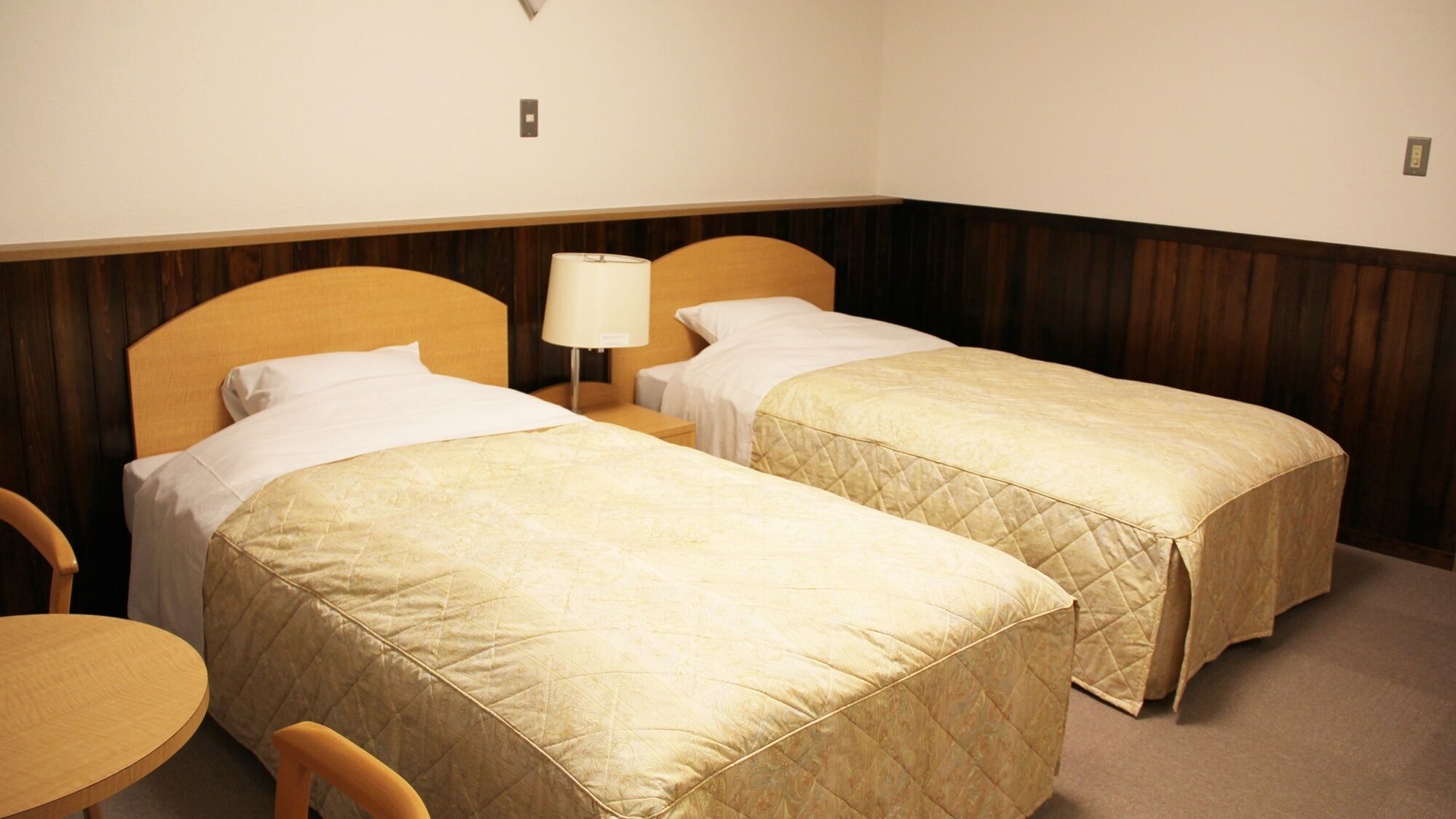 Una habitación de hotel con dos camas individuales, una mesita de noche con lámpara, una silla y una pequeña mesa redonda.