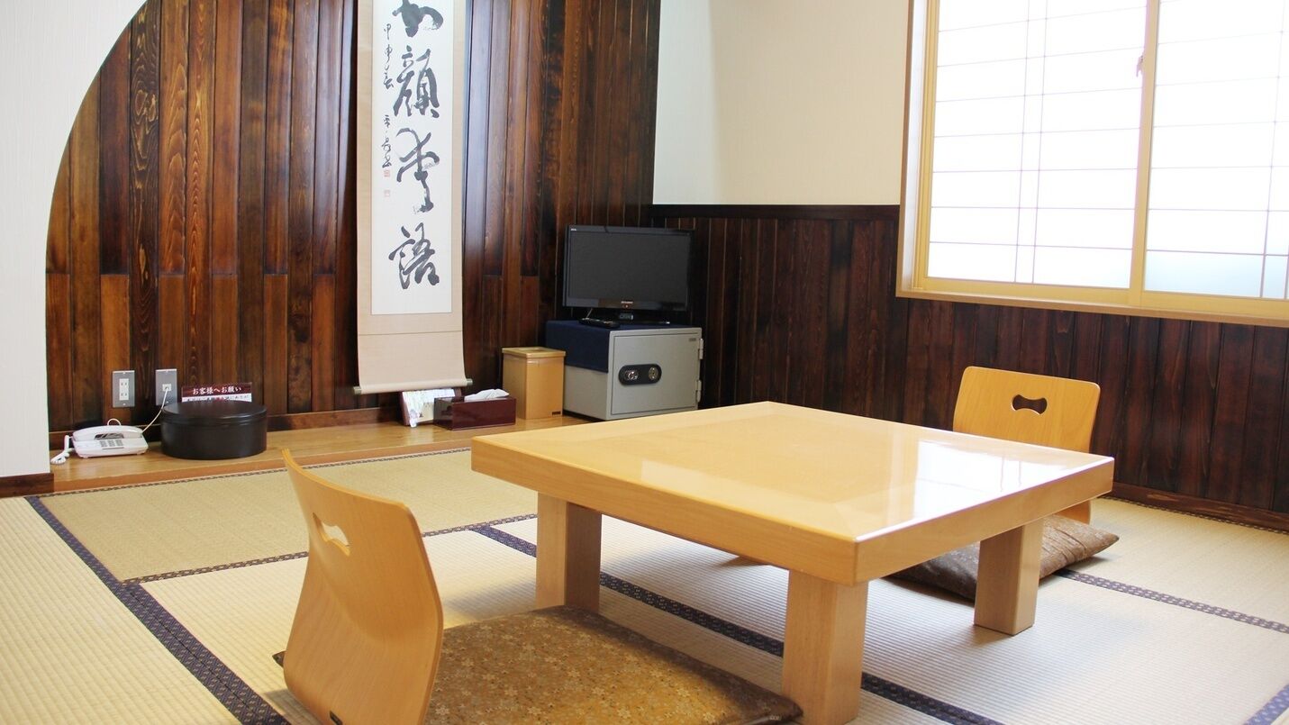 Una habitación japonesa tradicional con suelo de tatami, una mesa de madera, un televisor y un gran pergamino de caligrafía.